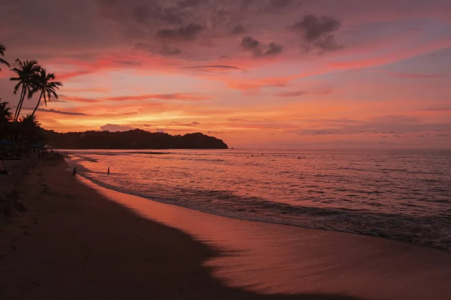 A surfer carrying a board walks along a sandy beach in Sayulita, Mexico, with palm trees and the ocean in warm sunset li
